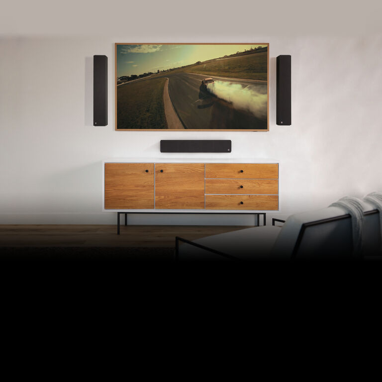 A living room with floor standing speakers, a subwoofer, and a wall mounted speaker all surrounding an electric fireplace.